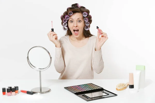 Mujer divertida con rizadores en el pelo pintando labios con lápiz labial, sentada a la mesa aplicando maquillaje con cosméticos decorativos faciales establecidos aislados sobre fondo blanco. Belleza mujer moda estilo de vida concepto . — Foto de Stock