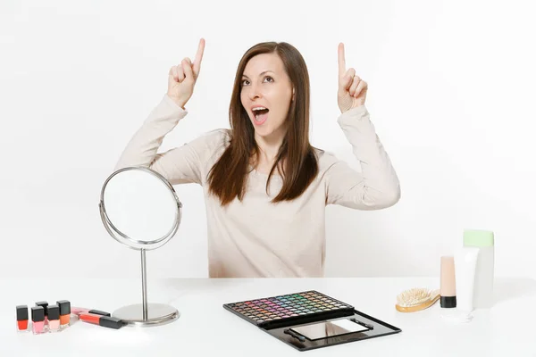 Preciosa mujer joven señalando con los dedos hacia arriba en el espacio de copia, sentado a la mesa aplicando maquillaje con cosméticos decorativos faciales establecidos aislados sobre fondo blanco. Belleza mujer moda estilo de vida concepto . — Foto de Stock