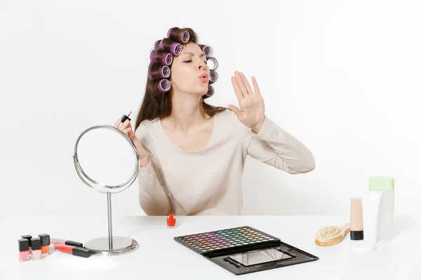 Mujer divertida con rizadores pintando uñas con esmalte de uñas rojo, sentada a la mesa aplicando maquillaje con cosméticos decorativos faciales establecidos aislados sobre fondo blanco. Belleza mujer moda estilo de vida concepto . — Foto de Stock