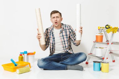 Confused sad shocked tired man with crossed rolls of wallpaper, sitting on floor with instruments for renovation apartment isolated on white background. Gluing accessories, tools. Repair home concept. clipart