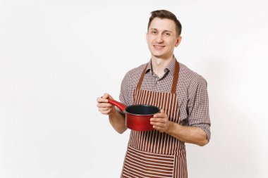 Young smiling man chef or waiter in striped brown apron, shirt holding red empty stewpan, pan or pot isolated on white background. Male housekeeper or houseworker. Kitchenware and cuisine concept. clipart