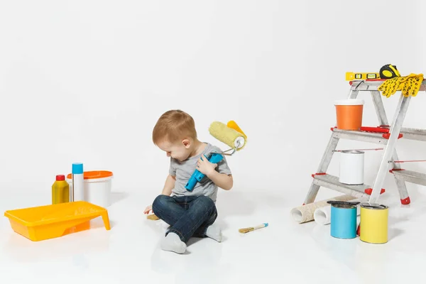 Pequeño niño lindo sentado en el suelo con instrumentos para la renovación habitación del apartamento aislado sobre fondo blanco. Fondo de pantalla, pegar accesorios, herramientas de pintura. Reparar casa. La paternidad, concepto de infancia . — Foto de Stock