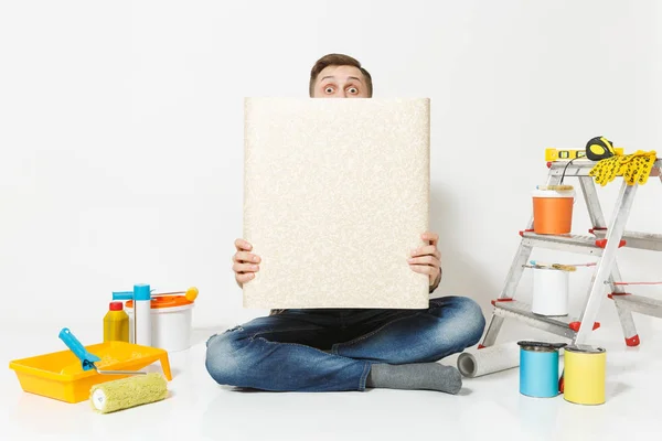 Schöner Mann sitzt auf dem Boden mit ausgeklappten Rolle von Tapeten Instrumente für die Renovierung Wohnung Zimmer isoliert auf weißem Hintergrund. Kleben Zubehör Malwerkzeuge. Reparaturheimkonzept. Kopierraum — Stockfoto