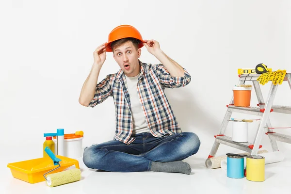 Διασκέδαση τρελός άνθρωπος στο πορτοκαλί προστατευτικό κράνος που κάθεται στο πάτωμα με τα όργανα για διαμέρισμα ανακαίνιση που απομονώνονται σε λευκό φόντο. Ταπετσαρία, κόλλημα αξεσουάρ, εργαλεία βαφής. Επισκευή concept home. — Φωτογραφία Αρχείου