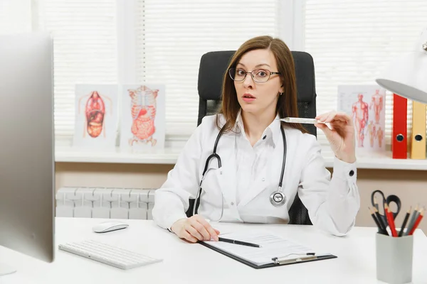 Female doctor sitting at desk with medical documents, clinical thermometer, high fever temperature in light office in hospital. Woman in medical gown, stethoscope in consulting room. Medicine concept.