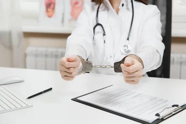Tutuklanan kadın doktor hastanede ışık ofiste tıbbi belgelerle masa başında oturan kapatın. Kadın tıbbi elbise, stetoskop, eller Oda danışmanlık içinde kelepçe ile. Tıp, hukuk kavramı.