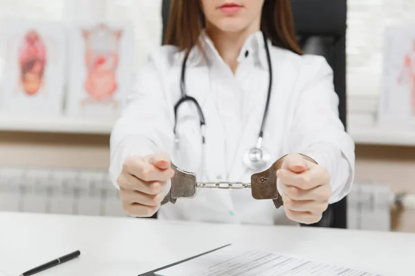 Närbild av arresterade kvinnliga läkare sitter vid skrivbord med medicinska handlingar i ljus kontor i sjukhuset. Kvinna i medicinsk klänning, stetoskop, händer med handbojor i samråd rum. Medicin, begrepp. — Stockfoto
