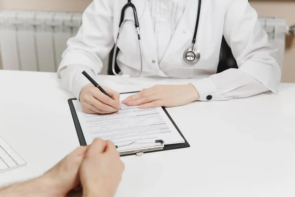 Jovem mulher sentada na mesa, trabalhando, preenchendo documentos médicos em escritório leve no hospital. Médica em vestido médico, estetoscópio em consultório com paciente. Conceito de medicina de saúde — Fotografia de Stock