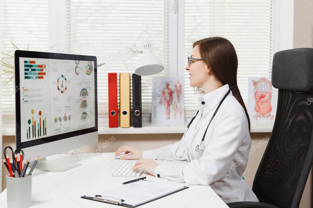 Young woman sitting at desk, working on modern computer with medical documents in light office in hospital. Female doctor in medical gown, stethoscope in consulting room. Healthcare, medicine concept.
