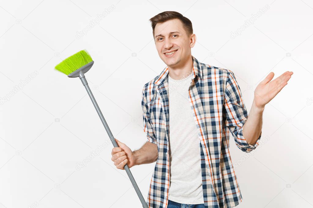 Young smiling happy housekeeper man in checkered shirt holding and sweeping with green broom isolated on white background. Male doing house chores. Copy space for advertisement. Cleanliness concept.