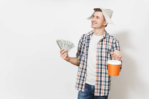 Jeune homme souriant au chapeau de journal tenant un paquet de dollars, de l'argent comptant et un seau de peinture vide avec un espace de copie isolé sur fond blanc. Instruments pour salle de rénovation. Réparation concept de maison . — Photo