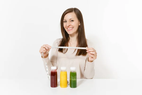 Mujer joven a la mesa con batidos desintoxicantes verdes, rojos y amarillos en botellas, cinta métrica aislada sobre fondo blanco. Nutrición adecuada, bebida vegetariana, estilo de vida saludable, concepto de dieta. Copiar espacio . — Foto de Stock