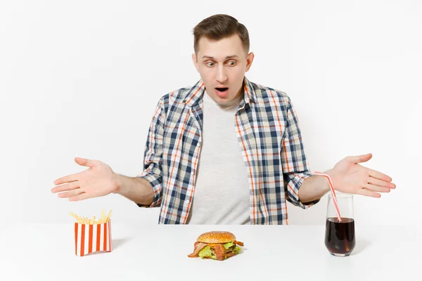 Schockierter junger Mann breitet Hände aus, sitzt am Tisch mit Burger, Pommes, Cola im Glas isoliert auf weißem Hintergrund. richtige Ernährung oder klassisches amerikanisches Fast Food. Werbefläche, Kopierfläche. — Stockfoto