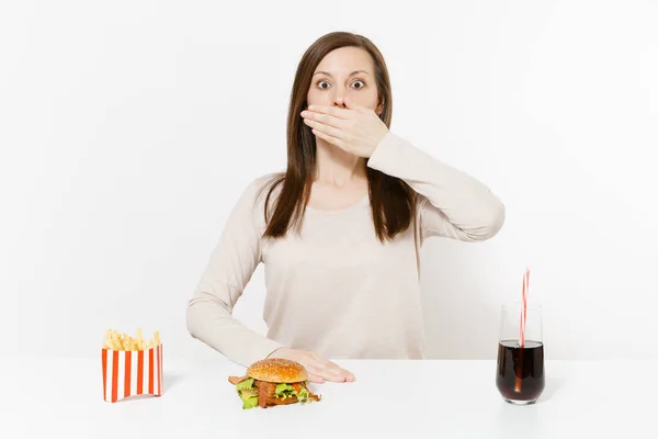 Frau bedeckt Mund mit der Hand am Tisch mit Burger, Pommes, Cola in Glasflasche isoliert auf weißem Hintergrund. richtige Ernährung oder klassisches amerikanisches Fast Food. Werbefläche mit Kopierfläche. — Stockfoto