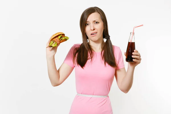 Sad young woman with tails, measuring tape at waist holding burger, cola in glass bottle isolated on white background. Proper nutrition or American classic fast food. Advertising area with copy space. — Stock Photo, Image
