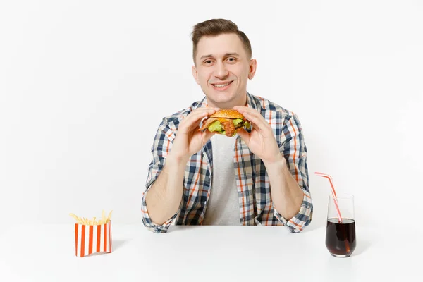 Bonito jovem sorridente sentado à mesa com hambúrguer, batatas fritas, cola em vidro isolado no fundo branco. Nutrição adequada ou fast food clássico americano. Área de publicidade com espaço de cópia . — Fotografia de Stock