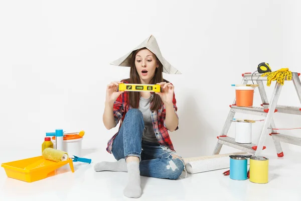 Mujer en sombrero de periódico sentado en el suelo con el edificio burbuja nivel de espíritu, instrumentos para la renovación apartamento aislado sobre fondo blanco. Fondo de pantalla, pegar accesorios, herramientas. Reparación concepto hogar . — Foto de Stock