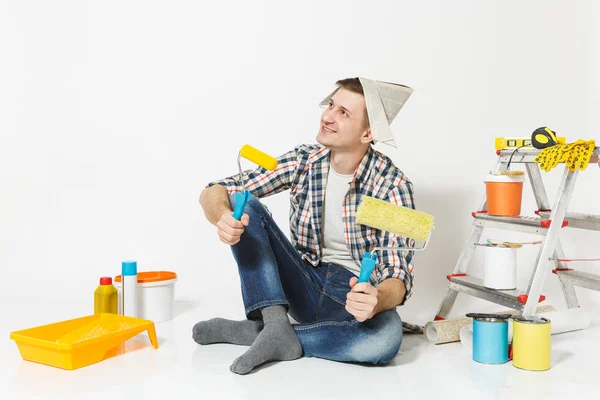 Ο άνθρωπος με καπέλο εφημερίδα κάθεται στο πάτωμα με κυλίνδρους χρωμάτων, μέσα για την ανακαίνιση Διαμέρισμα Δωμάτιο απομονωθεί σε λευκό φόντο. Ταπετσαρία, κόλλημα αξεσουάρ, εργαλεία βαφής. Επισκευή concept home. — Φωτογραφία Αρχείου