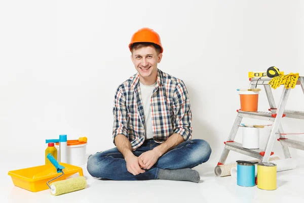 Homem sorridente em capacete de proteção laranja sentado no chão com instrumentos para renovação apartamento isolado no fundo branco. Papel de parede, acessórios de colagem, ferramentas de pintura. Reparação home concept . — Fotografia de Stock