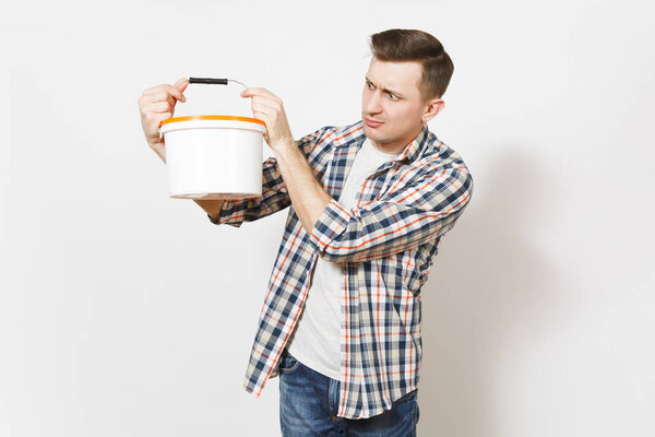 Young concerned handsome man in casual clothes holding and looking on empty paint bucket with copy space isolated on white background. Instruments for renovation apartment room. Repair home concept.