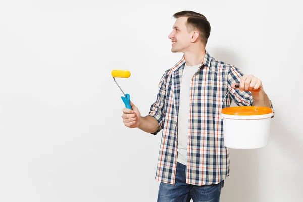 Joven hombre sonriente sosteniendo rodillo de pintura para la pintura de pared, cubo de pintura vacío y mirando a un lado en el espacio de copia aislado sobre fondo blanco. Herramientas para la renovación de la habitación del apartamento. Reparación concepto hogar . — Foto de Stock