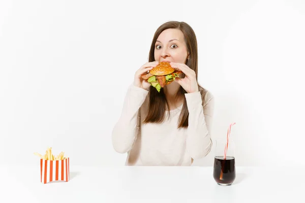 Aç genç kadın iştahla hamburger yiyor, tablo ile kızarmış patates, beyaz arka plan üzerinde izole şişede cola oturur. Doğru beslenme veya Amerikan Klasik fast food. Alan kopya alanı ile reklam. — Stok fotoğraf