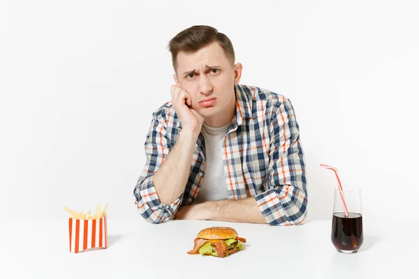 Szép szomorú ideges fiatal ember ül az asztal, hamburger, sült krumpli, cola pohár elszigetelt fehér background. Megfelelő táplálkozás, vagy a klasszikus amerikai gyorsétterem. Hirdetés terület másol hely. — Stock Fotó