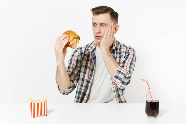 Knappe geschokt jonge man zit aan tafel met Hamburger, frietjes, cola in het glas geïsoleerd op een witte achtergrond. Goede voeding of Amerikaanse klassieke fastfood. Gebied met kopie ruimte reclame. — Stockfoto