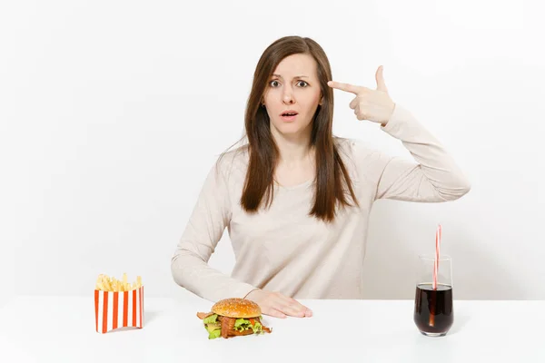 Donna triste punta dito a testa come se si sparasse a tavola con hamburger, patatine fritte, cola in bottiglia di vetro isolata su fondo bianco. Una corretta alimentazione o fast food americano. Con spazio di copia . — Foto Stock