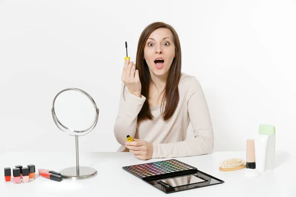 Atractiva joven mujer pintando pestañas con rímel, sentada a la mesa aplicando maquillaje con cosméticos decorativos faciales establecidos aislados sobre fondo blanco. Belleza mujer moda estilo de vida concepto . — Foto de Stock