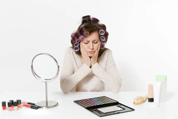 Mujer joven triste con rizadores en el pelo sentado en la mesa aplicando maquillaje con cosméticos decorativos faciales conjunto aislado sobre fondo blanco. Belleza concepto de estilo de vida femenino. Área con espacio de copia . — Foto de Stock