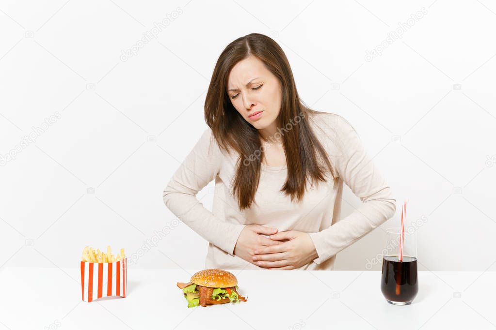 Illness woman put hands on pain abdomen, stomach-ache at table with burger, french fries, cola in glass bottle isolated on white background. Proper nutrition or American classic fast food. Copy space.