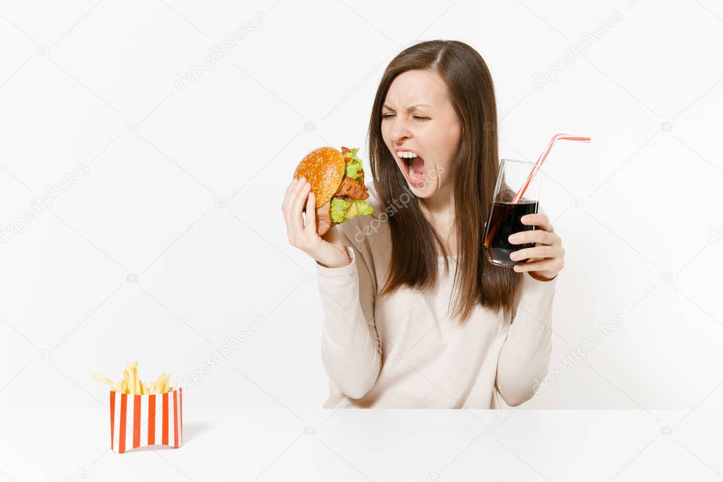 Angry screaming woman sitting at table with burger, french fries, cola in glass bottle isolated on white background. Proper nutrition or American classic fast food. Advertising area with copy space.