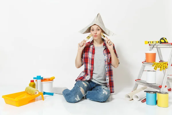 Mulher divertida em chapéu de jornal sentado no chão com escova, instrumentos para renovação apartamento quarto isolado no fundo branco. Papel de parede, acessórios para colar, ferramentas de pintura. Reparação home concept . — Fotografia de Stock