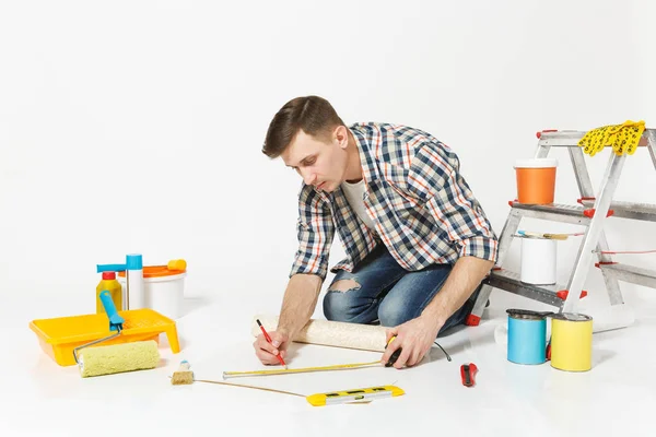 젊은 남자 롤 벽지, 측정 테이프, 연필, 재건축 아파트 룸을 위한 계기의 바닥에 앉아 흰색 배경에 고립. 액세서리, 도구 그림 붙이기 수리 홈 개념 — 스톡 사진