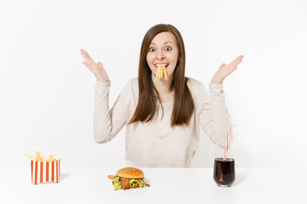 Mujer divertida en la mesa con varios pedazos de papas en la boca, papas fritas, hamburguesa, cola en botella de vidrio aislado sobre fondo blanco. Nutrición adecuada o comida rápida americana. Área con espacio de copia . —  Fotos de Stock