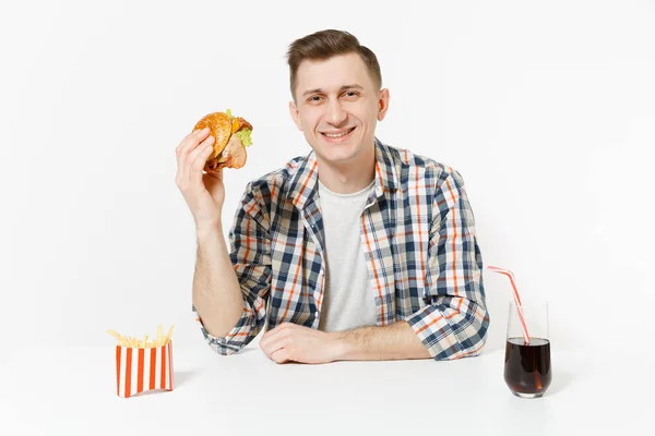 Bonito jovem sorridente sentado à mesa com hambúrguer, batatas fritas, cola em vidro isolado no fundo branco. Nutrição adequada ou fast food clássico americano. Área de publicidade com espaço de cópia . — Fotografia de Stock