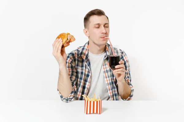 Knappe jonge man zit aan tafel met Hamburger, frietjes, cola drinken uit glas geïsoleerd op een witte achtergrond. Goede voeding of Amerikaanse klassieke fastfood. Gebied met kopie ruimte reclame. — Stockfoto