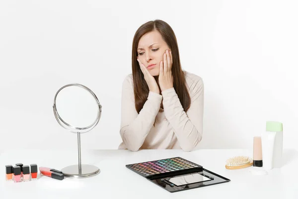 Sorprendida joven molesta sentada a la mesa aplicando maquillaje con cosméticos decorativos faciales establecidos aislados sobre fondo blanco. Belleza concepto de estilo de vida femenino. Área de publicidad con espacio de copia . — Foto de Stock