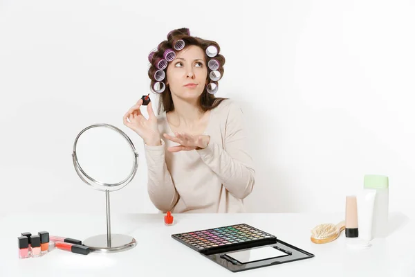 Mujer divertida con rizadores pintando uñas con esmalte de uñas rojo, sentada a la mesa aplicando maquillaje con cosméticos decorativos faciales establecidos aislados sobre fondo blanco. Belleza mujer moda estilo de vida concepto . — Foto de Stock