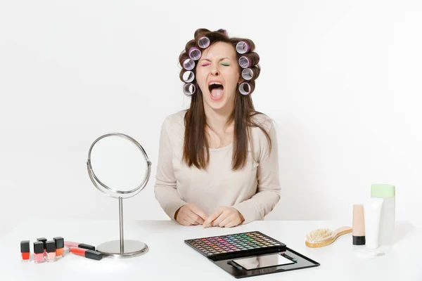 Fun crying woman with curlers on hair sitting at table applying makeup with set facial decorative cosmetics isolated on white background. Beauty female fashion lifestyle concept. Area with copy space. — Stock Photo, Image