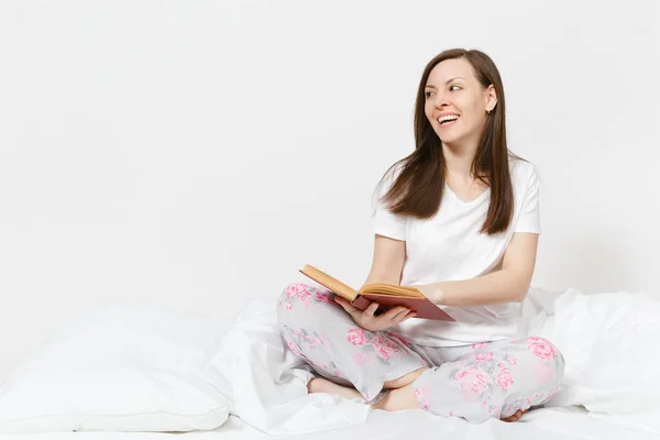 Joven mujer feliz sentada en la cama con sábana blanca, almohada, manta sobre fondo blanco. Belleza femenina pasar tiempo en la habitación, leer libros, disfrutar del descanso. Relájate, concepto de buen humor. Lugar para el texto . — Foto de Stock