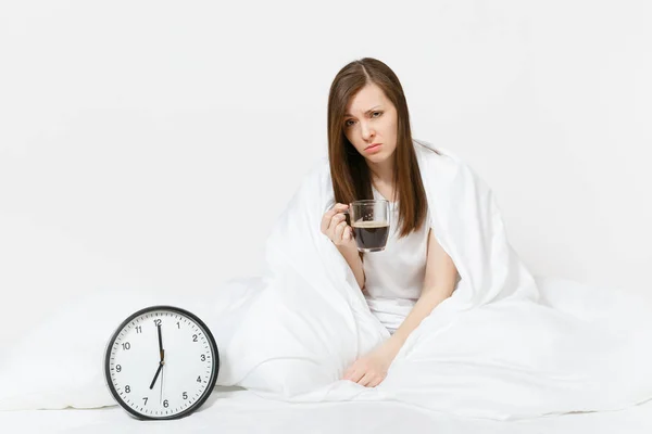 Tired sad woman sitting in bed with cup of coffee, round clock, white sheet, pillow, wrapping in blanket on white background. Female wake up early in morning, time in room. Rest, good mood concept.