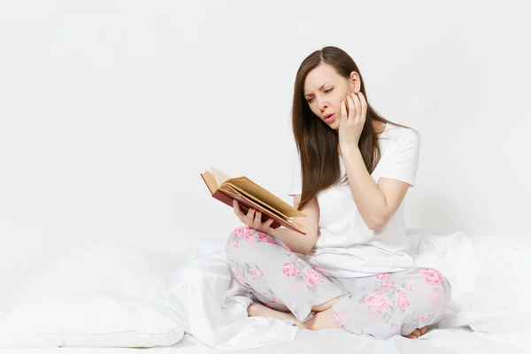 Ung lugn kvinna sitter i sängen med vitt lakan, kudde, filt på vit bakgrund. Skönhet kvinna umgås i rummet, läsa bok, njuta av resten. Koppla av, gott humör koncept. Plats för text. — Stockfoto
