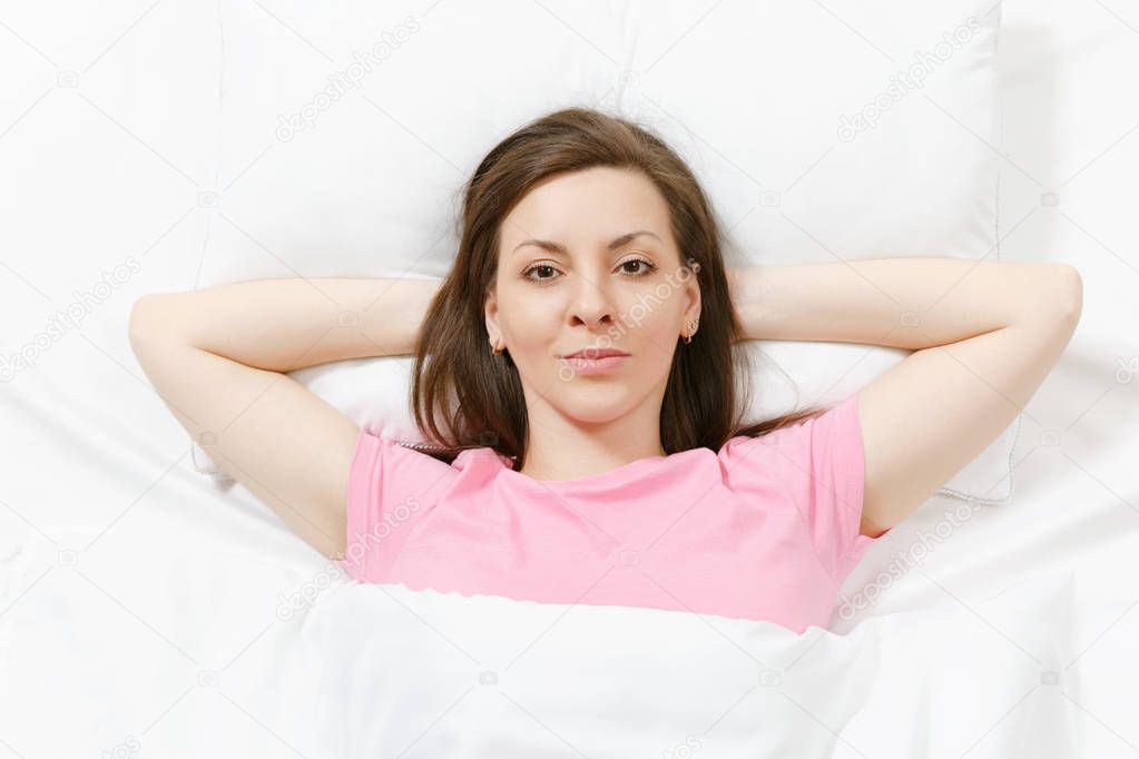 Top view of happy brunette young woman lying in bed with white sheet, pillow, blanket, hands under head. Smiling female spending time in room. Rest, relax, good mood concept. Copy space advertisement.