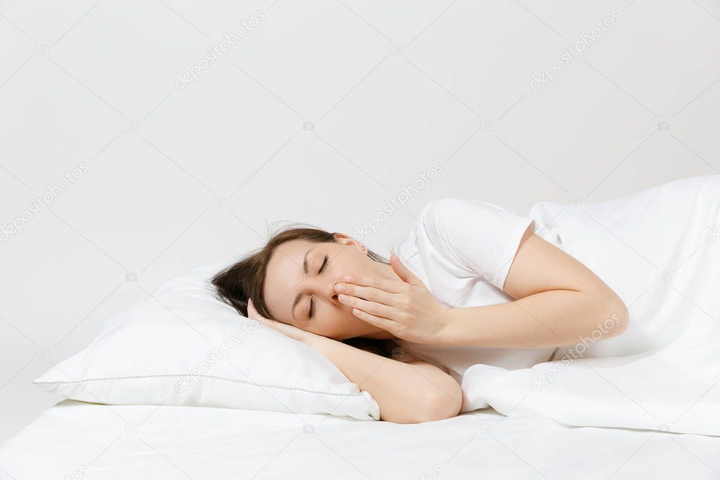 Calm young brunette woman lying in bed with white sheet, pillow, blanket on white background. Yawning beauty female spending time in room. Rest, relax, good mood concept. Copy space for advertisement.