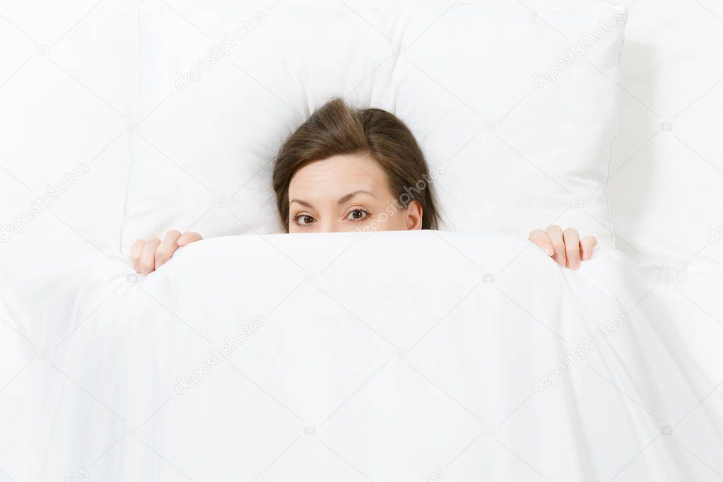 Top view of head of happy brunette young woman lying in bed with white sheet, pillow, blanket, peek out. Female spending time in room. Rest, relax, good mood concept. Copy space for advertisement.