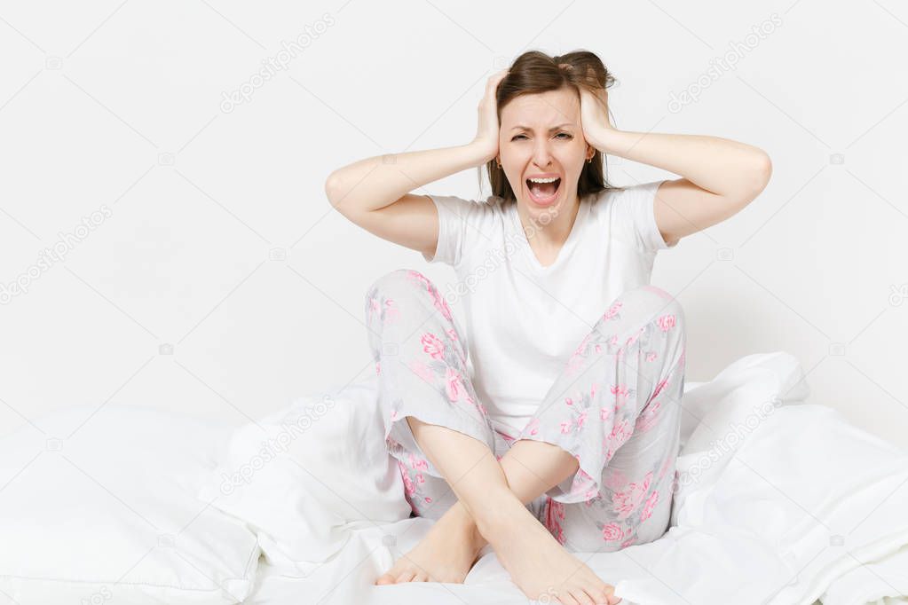 Stressed crazy mad young screaming woman sitting in bed with white sheet, pillow, blanket. Shocked female cover ears with hands, spending time in room. Bad mood, stress, problem concept. Copy space.