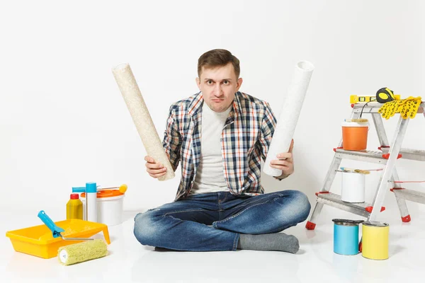 Confundido hombre molesto cansado con rollos de papel pintado, sentado en el suelo con instrumentos para la renovación apartamento aislado sobre fondo blanco. Fondo de pantalla, pegar accesorios, herramientas. Reparación concepto hogar . — Foto de Stock