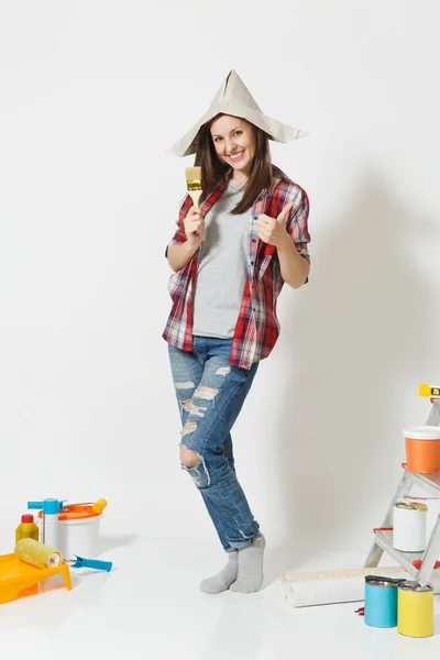 Full length portrait of woman in newspaper hat stands with brush, instruments for renovation apartment home isolated on white background. Wallpaper, gluing accessories, painting tools. Repair concept.